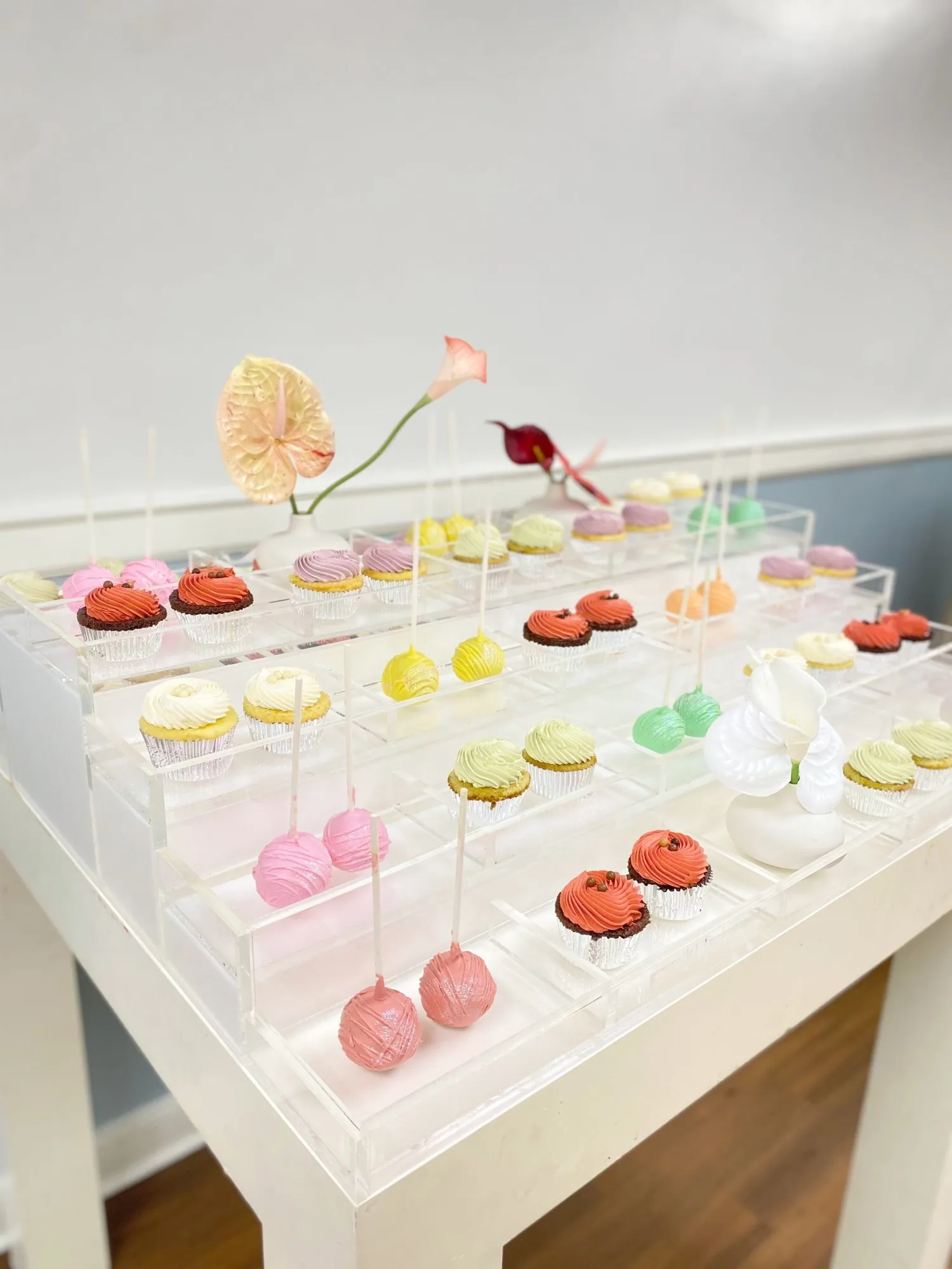 Pastel dessert table on tiered acrylic risers with cake pops, mini cakes, and bite-size desserts, styled with tropical flowers