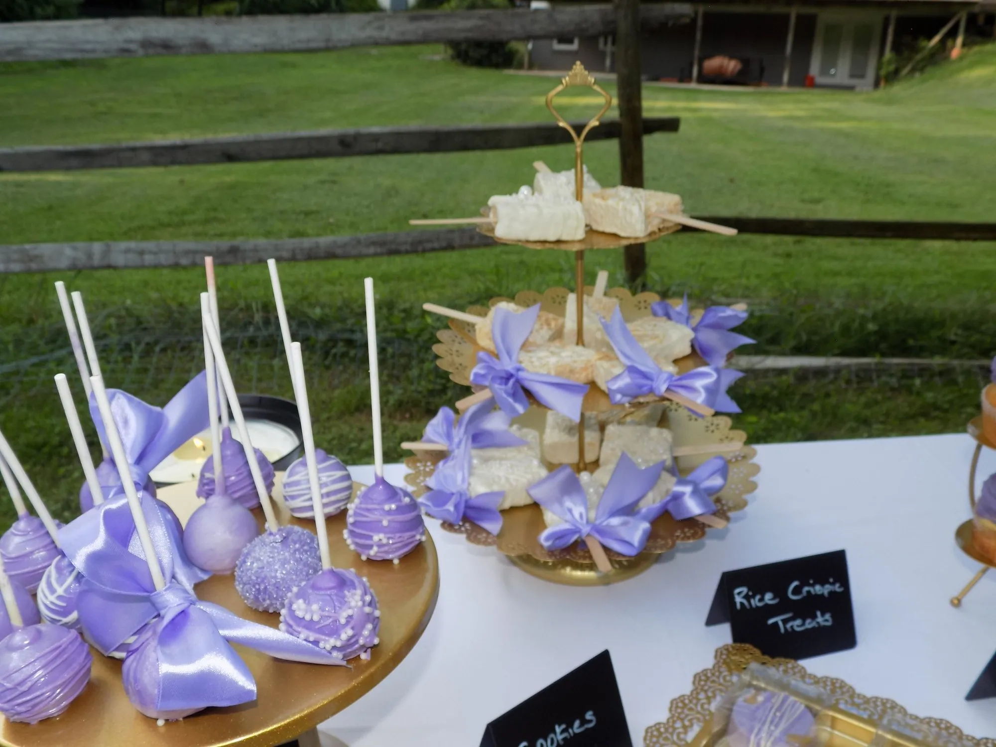 Outdoor dessert table with lavender cake pops tied with ribbon bows, gold stands, and dessert trays