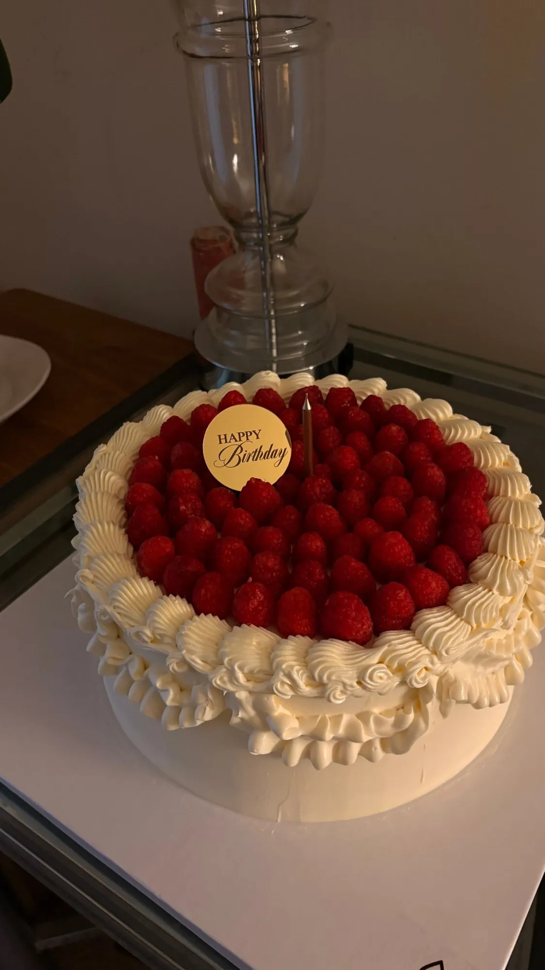Round white cake with ruffled buttercream edge, topped with fresh raspberries and a gold "Happy Birthday" tag